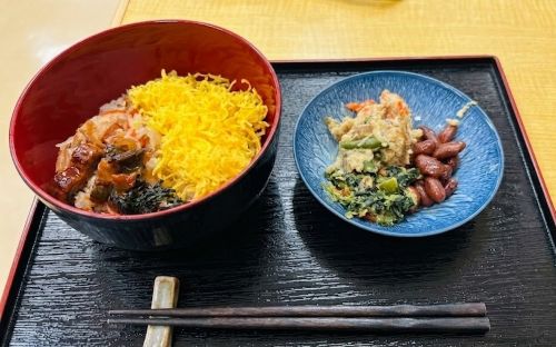 お盆に配膳された行事食(うなぎ入り散らし寿司と副菜)の盛り付け写真
